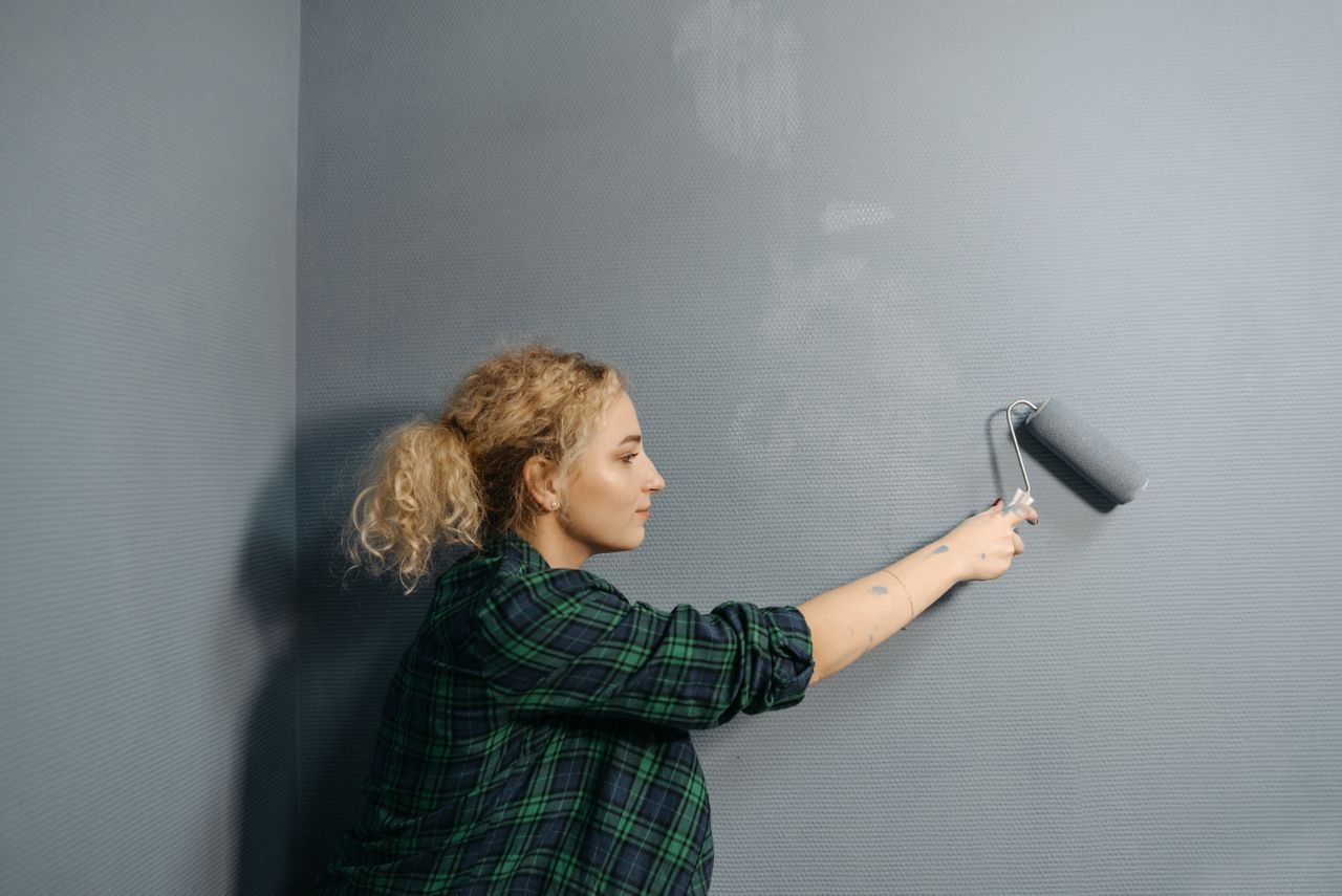 Free Young woman uses a paint roller to apply grey paint on an indoor wall, renovating her space. Stock Photo